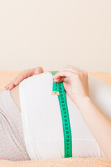 Young beautiful pregnant girl measuring her belly with a tape in home on bed. Healthy pregnancy concept