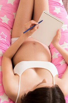 Pregnant woman writing baby names list on sofa, closeup. Pregnant mom writes in a notebook and to-do list shopping