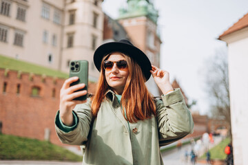 Attractive young female tourist is exploring new city. Redhead girl holding a smartphone in Krakow. Traveling. Happy optimistic girl walking in city and makes selfie poses against historic building