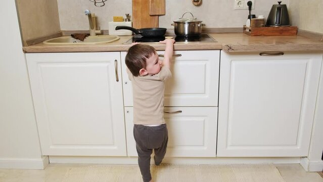 Toddler baby climbs on a hot electric stove in the home kitchen. A small child touches the surface of the stove with his hand at the risk of getting burned. Kid aged one year eight months