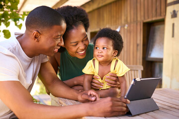 Family With Young Daughter Sitting On Table Outdoors At Home Making Video Call On Digital Tablet