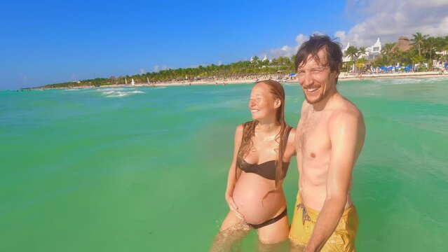 A slow-motion stock video captures a pregnant woman and a man joyfully exploring the tropical sea. They walk hand in hand, capturing their holiday memories with a selfie video using an action camera.