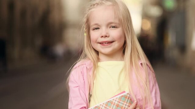 Portrait of cute little blond girl with backpack hold notebooks and looking at the camera standing on city street alone Adorable kid enjoying free time after lessons outdoors