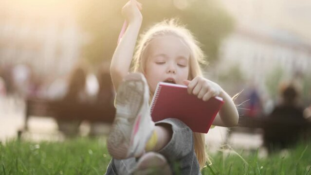 Cute little blond girl thinks questioningly and finding solution idea while write homework make notes sitting on grass at park Pretty child kid has remote distance education outdoors