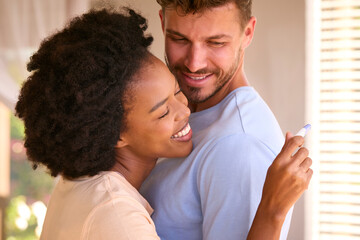 Hugging Multi-Racial Couple In Bedroom At Home Celebrating Positive Pregnancy Test Result