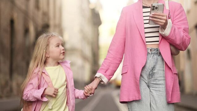 Cute little blond girl with backpack get back to home after study lessons from school holding her mother's hand and talking to her while mom ignoring daughter using smartphone at city street 