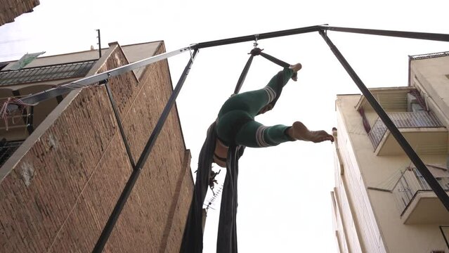 Woman hanging on aerial fabrics in an inner courtyard of buildings, training circus and show in her house, back with tattoos and head with dreadlocks.