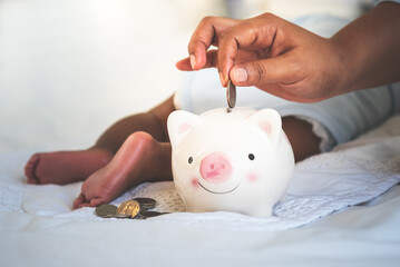 Mother is putting money in a piggy bank, while her baby newborn is sleeping on white bed, concept...