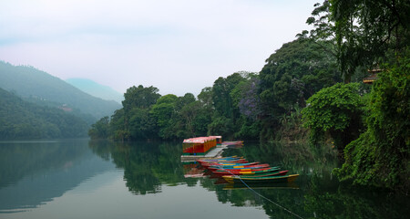In this picturesque landscape photo, Phewa Lake in Pokhara, Nepal, unfolds its serene beauty. The calm waters of the lake reflect the surrounding mountains, creating a breathtaking panorama. A cluster