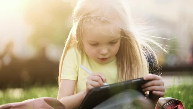 Charming little girl child drawing sketch from nature using tablet at park Pretty kid with long blond hair painting by stylus at screen of digital pad outdoors 