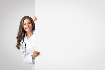Smiling senior woman medical worker looking out behing white advertising board with free space for text or design