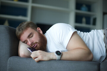 Young bearded man sleep at home on couch with his cell mobile phone in living room. Smartphone...