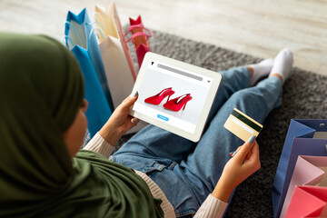 Young arabic woman in hijab using credit card and tablet with fashion store on screen, offering...