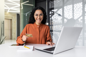 Portrait of a young Hispanic woman working in the office using a laptop, holding a credit card and smiling at the camera.