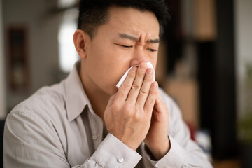 Cold, flu, allergy concept. Sick middle aged asian man sneezing in napkin, ill male feeling unwell, sitting on couch