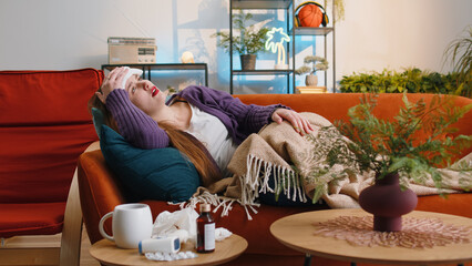 Portrait of ill woman suffering from cold or allergy pain lying on couch in living room at home. Sick girl with towel on head blows sneezes wipes snot into napkin. Coronavirus quarantine, pandemic