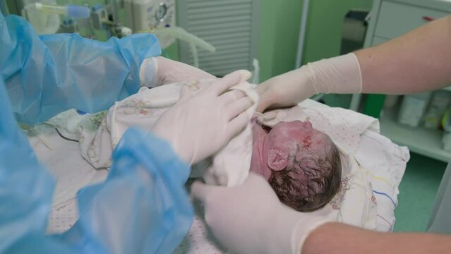 The gloved hands of a doctor and a nurse wipe a newborn baby from the remains of fetal blood and protective film. Gynecology. Maternity ward.