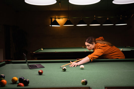 Beautiful girl learning to play billiards watching online video lesson of playing billiards using laptop on pool table