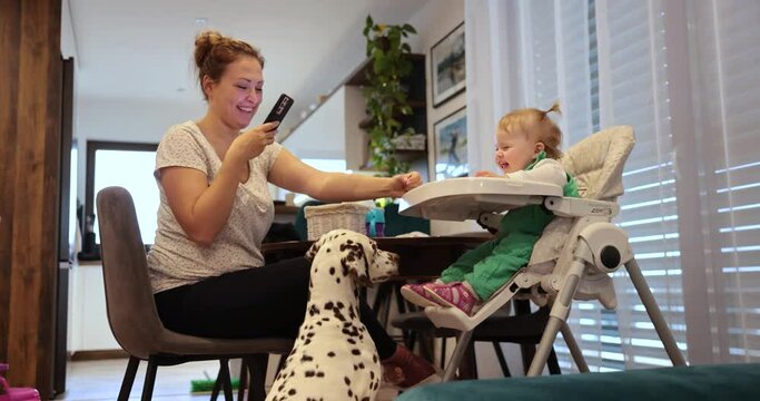 Mom feeding her one year old daughter with dalmatian dog around. Recording and taking pictures on phone. Having a nice time. Laughing smiling eating. Baby in green dress. Modern home, kitchen.