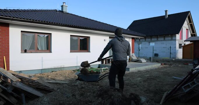 Good looking man worker throwing earth and mold in to garden wheelbarrow and riding it. Beautifull surrounding nature and high snowy mountains. Cold windy weather. Working around house in garden 4k 
