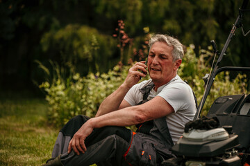 A mature man is taking a break and talking on the phone after working on a lawnmower around a lawn in the backyard.