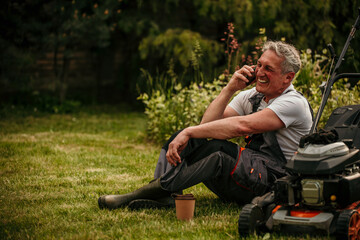 A mature man is taking a break and talking on the phone after working on a lawnmower around a lawn...