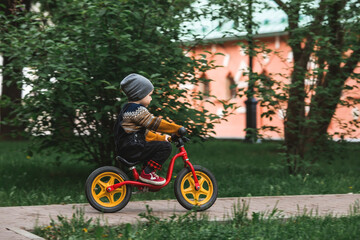 A cheerful little boy rides a bicycle outdoors. A happy child walks in the spring park. The kid is dressed in a fashionable sweater and denim overalls.