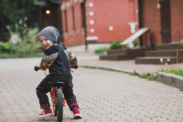 A cheerful little boy rides a bicycle outdoors. A happy child walks in the spring park. The kid is dressed in a fashionable sweater and denim overalls.