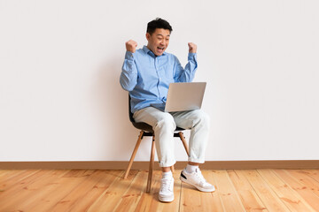 Overjoyed asian middle aged man sitting on chair with laptop and making YES gesture, joyful male celebrating success