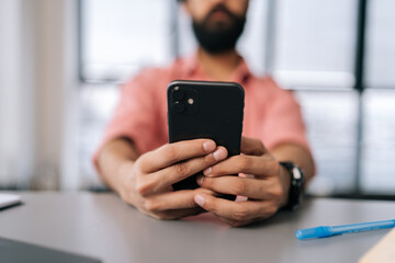 Close-up cropped selective focus shot of unrecognizable bearded man using smartphone sitting at office desk with book and work documents by window. Freelancer male texting message on mobile phone.