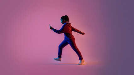 Black lady in sportswear walking and using cellphone over pink neon background, full body, side view, free space