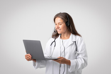 Cheerful european senior woman doctor in white coat with stethoscope, headphones video calling on laptop