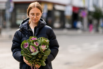 Fototapeta premium Smiling young woman standing in the street and gently holding large bouquet of beautiful ornamental cabbage brassica in hands