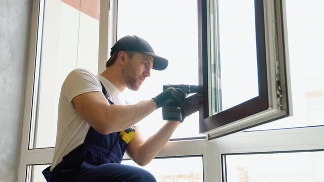 Workman in overalls installing pvc window with screwdriver