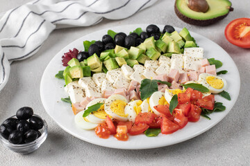Healthy cobb salad with ham, feta cheese, avocado, tomato, black olives and eggs on white plate on gray table. Ketogenic lunch