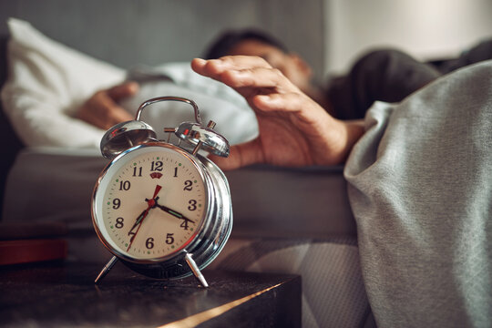 Alarm clock, wake up and man sleeping in the bedroom of his modern apartment in the morning. Lazy, resting and hand of male person taking a nap and dreaming in bed with time watch at his home.