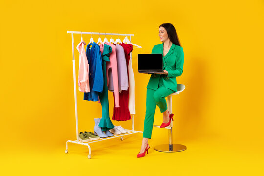 Lady stylist showing laptop with blank screen, advertising store website near clothing rail with trendy clothes, mockup