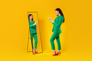 Trendy lady pointing at her reflection in mirror posing after successful shopping, wearing green jacket trouser suit