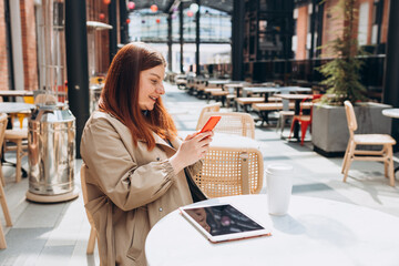 Young woman enjoying a coffee, sitting on the cafe terrace on the modern city street. Person sitting at table and using smartphone outdoors. Online education, order, working or shopping concept