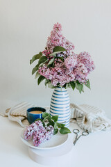 a bouquet of lilacs in a vase on a white table, a cup of coffee and scissors nearby