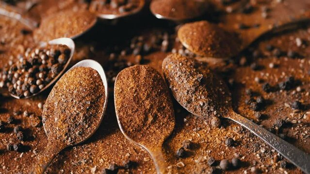 Spoons with Indian Spices Rotate on a Colorful, Wooden Background. Close up. Slow motion. Scattered spice mix, grains black peppercorns, turmeric, paprika, coriander, oregano. Backgrounds, texture.