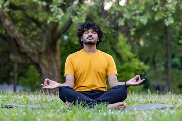 Young hispanic man relaxing in park, meditating and practicing yoga in lotus pose, sitting on grass on fitness mat with eyes closed