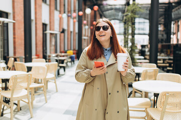 Happy cheerful redhead woman in fashion office clothes and sunglasess posing with eco cup of tea. Woman holding smartphone and bio coffee cup. Urban lifestyle concept