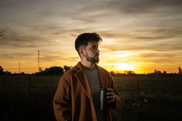 young man drinking coffee in the field with a sunset in the background