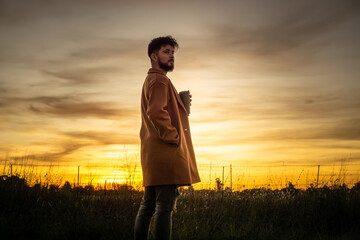 young man drinking coffee in the field with a sunset in the background