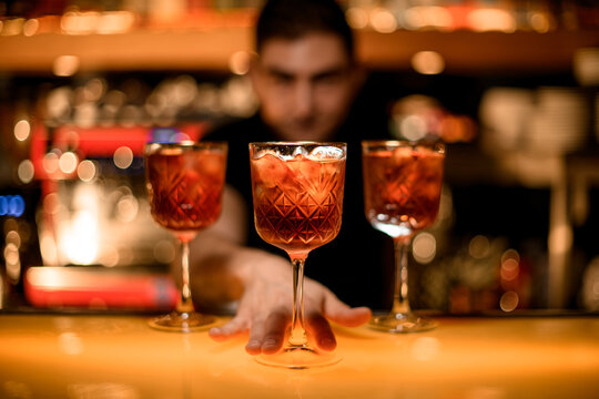 Hand of bartender serving cold red cocktails with ice cubes in glasses on the bar counter in the bar