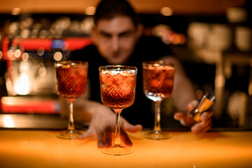 Male bartender gives to cameraman cold red cocktail with ice cubes in glass on the bar counter in the bar