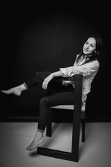 emotional portrait of cute and seductive brunette girl sitting in pants and white shirt on chair isolated on dark background