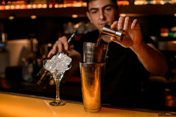 beautiful view of steel shaker cups on bar and bartender pouring drink from jigger into it