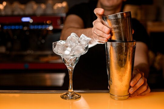 Bartender holds steel shaker cups in hands. Martini glass with ice cubes stands nearby